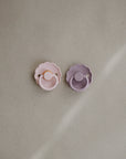 The FRIGG Daisy Silicone Pacifier 2-Pack features one pink and one lavender pacifier with scalloped edges, made from medical-grade silicone for comfort—shown side by side on a neutral, textured background.