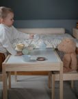 A young child with the mushie Silicone Pacifier Clip | Hera pours imaginary tea for a plush teddy bear at a small table, hosting a pretend tea party in a cozy, minimalist room.