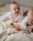 Two babies play with stacking cups on a mushie Organic Cotton Muslin Swaddle Blanket.
