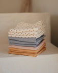 The mushie Organic Cotton Muslin Cloths 3-Pack features soft, neutral-colored cloths—white with a floral pattern, gray, and pink—neatly stacked on a textured white surface, ideal for nursery use.