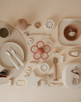Flat lay of mushie Lunchbox and other baby feeding items in neutral and pastel tones, including plates, bowls, utensils, sippy cup, pacifiers, bib, flower-shaped teether, and a star item—all neatly arranged on a beige background.