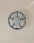A blue, round mushie Suction Spinner Toy, made from food-grade silicone and resembling a simple ship's wheel, attaches to a plain light-colored wall.