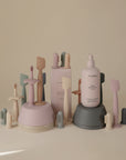 Pastel Mushie baby care items—bottles of lotion, silicone toothbrushes, and a Mushie Diaper Cream Applicator—arranged aesthetically on inverted bowls against a neutral background.
