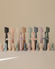 A row of pastel-colored Mushie silicone baby toothbrushes, finger brushes, and a Diaper Cream Applicator with matching stands are neatly arranged against a beige background.