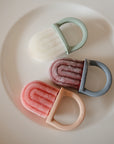 Three colorful rainbow-shaped popsicles with rounded green, blue, and peach handles rest on a white plate. Made using the mushie Toddler Ice Pop Tray, each pop features "mushie" embossed on the handle for a fun treat.