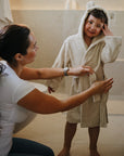 A woman helps a smiling child wearing the mushie Bear Robe in a bathroom.