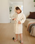 A toddler in a mushie Bear Robe ties the belt, standing barefoot in a cozy living room.
