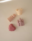 The mushie Princess Mold Free Bath Play Set includes four pastel food grade silicone bath toys—a yellow crown, a bear in a pink gown, a faceted pink heart, and a pink castle—for fun, mold-free play.