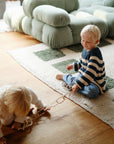 Two young children play on a wooden floor beside a green sofa. One smiles while holding mushie's Chain Link Rings, and the other crawls nearby—both engaging their fine motor skills with this colorful toy.