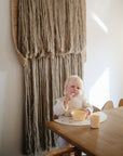 A young child with blonde hair eats at a wooden table from a mushie Round Dinnerware Bowl, BPA-free and yellow, with a matching cup nearby. Sunlight streams past a neutral woven wall tapestry in the background.