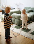 Two young children with blonde hair play barefoot in a living room with a green couch and patterned rug. One holds mushie Chain Link Rings, engaging in fun play that encourages fine motor skills.