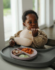 A young child in a high chair feeds independently with mushie Silicone Toddler Starter Spoons, using a divided plate and wearing a patterned shirt and bib in a bright, sunlit room.