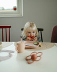 A young child with blonde hair eats watermelon at a table set with mushie Square Dinnerware Plates, a pink sippy cup, and a teething toy. The background features a window and simple chairs.
