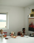 Four young children sit at a white dining table, eating from mushie Square Dinnerware Bowls while coloring. The bright room features natural light, a modern gold lamp overhead, and a colorful poster on the wall behind them.