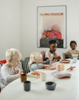 Four young children sit around a white table using mushie Square Dinnerware Bowls. The BPA-free plastic bowls, along with plates and cups, are set before them as natural light shines in and a colorful abstract poster decorates the background.