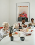 Four children sit at a white table eating watermelon, using mushie Round Dinnerware Plates and colorful cups. Sunlight fills the room, and a vibrant painting decorates the wall behind them.