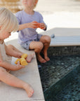 Two young children sit by a tiled pool, dipping their feet in the water. One plays with a yellow mushie Vehicles Mold Free Bath Play Set toy while the other holds an orange one. Both are relaxed, enjoying playtime together.