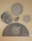 The mushie Lunchbox set features gray non-toxic plastic baby feeding items—divided plate, utensils, sippy cup, lidded container, bowl with spoon, and silicone placemat—arranged neatly on a beige surface.