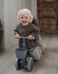 A laughing toddler rides a gray mushie Mini Toddler Bike down a hallway.