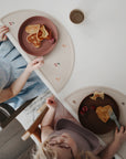 Two young children in light clothes sit at a white table with mushie Silicone Placemats, each holding a plate of heart-shaped pancakes and raspberries. A cup is placed next to each plate on the food-grade silicone mats.