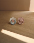 The FRIGG Daisy Silicone Pacifier 2-Pack by FRIGG features one blue and white, and one pink and mauve pacifier, each with a daisy-shaped shield and medical grade silicone nipple, displayed on a brown surface in soft natural light.