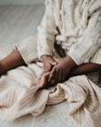 A child sits on a mushie Organic Cotton Muslin Swaddle Blanket, touching their bare foot.