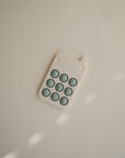 The mushie Phone Press Toy is a small, cream-colored sensory toy with rounded edges and nine blue-gray circular buttons, shown resting on a light, neutral surface with soft shadows.