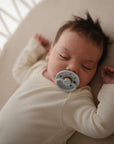 A dark-haired baby sleeps peacefully on a mushie Mini Muslin Crib Sheet, wearing a cream onesie and holding a gray pacifier, arms raised beside their head.