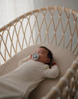 A baby in a cream-colored onesie sleeps in a light wooden bassinet fitted with the mushie Extra Soft Bassinet Muslin Crib Sheet, holding a pacifier as gentle daylight filters through the sheer curtain beside the crib.