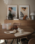 A wooden dining table is set with candles, baby mugs, a bib, and a mushie Bluey x Mushie Silicone Placemat. In the background, a sideboard with decor and two framed abstract art pieces rests against a beige wall.