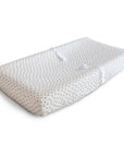 The mushie Extra Soft Muslin Changing Pad Cover in white features small, evenly spaced brown dots and comes with a rectangular pad and a safety strap for secure diaper changes.