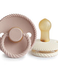 The FRIGG Rope Natural Rubber Pacifier 2-Pack by Mushie includes a dusty pink pacifier with a ring and a cream-colored one, both with textured rope-like edges and natural rubber latex nipples, and feature the embossed FRIGG brand name.