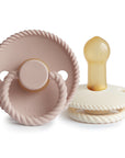 Two baby essentials are shown: the Mushie FRIGG Rope Natural Rubber Pacifier 2-Pack, featuring beige and white pacifiers with round, rope-like edges and ring handles. "FRIGG" branding is visible on one.