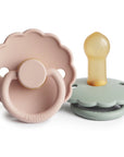 The FRIGG Daisy Natural Rubber Pacifier 2-Pack features two scalloped-edge silicone pacifiers: one pale pink daisy with a ring handle and one light green, both embossed with “FRIGG” and made from natural rubber latex.