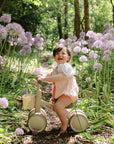 A smiling baby on a mushie Mini Toddler Bike rides among tall purple flowers on a sunny path.