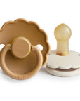 The FRIGG Daisy Natural Rubber Pacifier 2-Pack features two daisy-shaped silicone pacifiers with scalloped edges—one tan and upright, the other cream-colored on its side showing the natural rubber latex nipple.