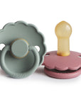 The FRIGG Daisy Natural Rubber Pacifier 2-Pack includes sage green and dusty rose pacifiers, each with a scalloped daisy-shaped shield, natural rubber latex nipple, and the FRIGG logo.