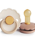 The FRIGG Daisy Natural Rubber Pacifier 2-Pack features two daisy-shaped pacifiers: one upright showing a natural rubber nipple with a brown base, and one lying flat with a beige front and brown ring handle. Both are branded FRIGG.
