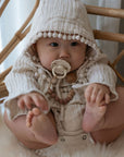 A baby in a textured cream hooded outfit with pom-pom trim sits on a fluffy surface, holding its feet and looking ahead, with a FRIGG Natural Rubber pacifier from the FRIGG 6-Pack clipped to its clothing.