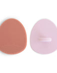 The mushie Cradle Cap Brushes in Rose/Soft Lilac are two oval silicone scrubbers—one side with brown bristles for gentle exfoliation, the smooth pink side features a handle and "mushie" embossed, perfect for baby bath time.