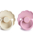 The FRIGG Daisy Natural Rubber Pacifier 2-Pack includes two daisy-shaped pacifiers with scalloped edges, in cream and light pink, round handles with the FRIGG logo, and natural rubber latex nipples for comfort.