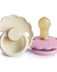 The FRIGG Daisy Natural Rubber Pacifier 2-Pack by FRIGG features a beige daisy-shaped pacifier with a ring handle and a pastel pink pacifier, both with natural rubber nipples, displayed upright on a white background.