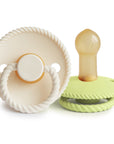 The Mushie FRIGG Rope Natural Rubber Pacifier 2-Pack includes two newborn essentials with rope-like edges: one beige with a ring handle and one light green with a natural rubber latex nipple, shown lying on its side.
