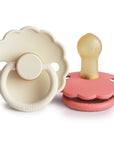 The FRIGG Daisy Natural Rubber Pacifier 2-Pack by FRIGG is shown in cream and pink. The cream pacifier is daisy-shaped with a scalloped edge and handle, while the pink one lies on its side, displaying its natural rubber latex nipple.