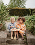 Two young children sit on stone steps outdoors in plaid shirts. One holds a tray of pastries while the other eats. A Mushie Wooden Puzzle adds an educational touch to the lush garden scene.