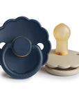 The FRIGG Daisy Natural Rubber Pacifier 2-Pack features two daisy-shaped pacifiers—one navy blue and one beige—both with scalloped edges and natural rubber latex nipples. The blue stands upright while the beige rests on its side.