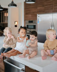 Four toddlers sit on a kitchen island with cups, cookies, and pacifiers. Nearby, FRIGG Andersen Fairytale Silicone Pacifier 2-Pack by FRIGG—made in Denmark—rests on the counter amid sleek appliances and wooden cabinets.