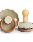 The FRIGG Daisy Natural Rubber Pacifier 2-Pack | Limited Edition features two beige and brown FRIGG pacifiers with scalloped edges—one stands upright, showing the latex nipple, and one lies flat with a round handle for baby comfort.