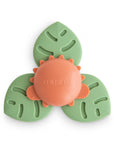 The mushie Suction Spinner Toy in Dino features three green leaf-shaped arms and an orange sun center embossed with "mushie," crafted from food-grade silicone for a delightful sensory play experience.