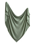 A mushie Stretchy Swaddle in light green hangs draped in a curved, symmetrical shape with soft folds and ruffled edges against a white background.